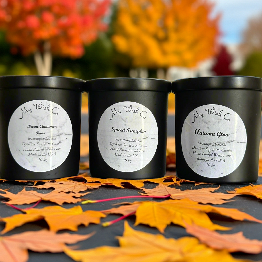 Three black candles labeled 'Woven Cinnamon', 'Spiced Pumpkin', and 'Autumn Glow' on a surface with autumn leaves.