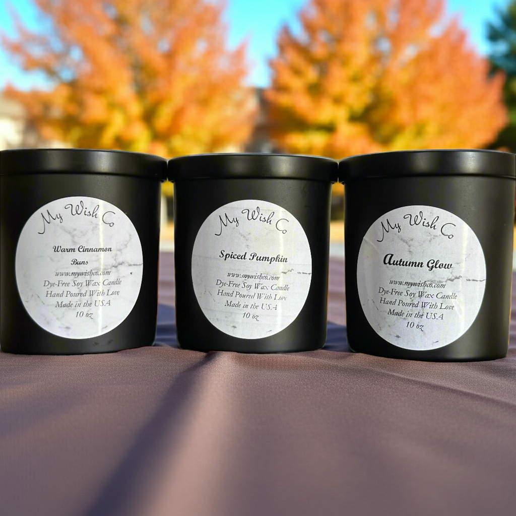 Three black candles with labels on a surface with autumn foliage in the background