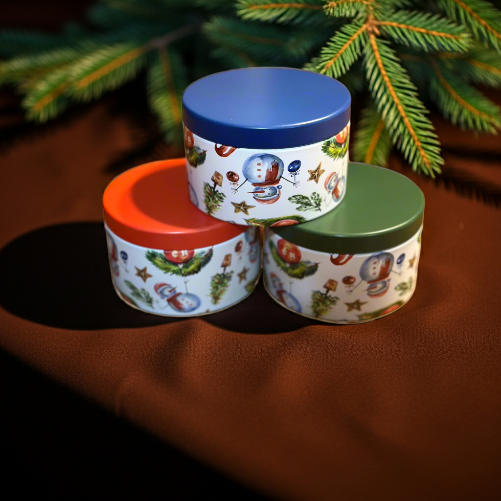 Three decorative tins with colorful lids stacked on a brown surface with a Christmas tree in the background.