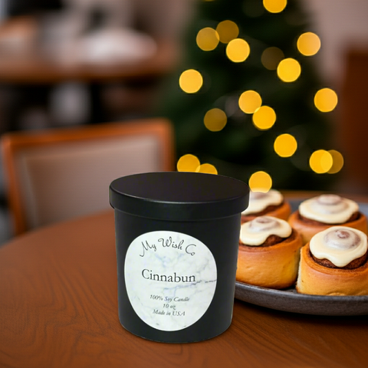 Black candle labeled 'Cinnabun' on a table with cinnamon buns and a blurred Christmas tree in the background.