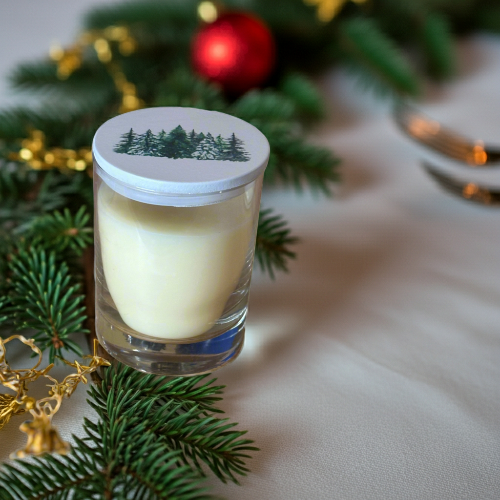 Glass candle with a lid featuring a forest design, surrounded by Christmas decorations on a table.