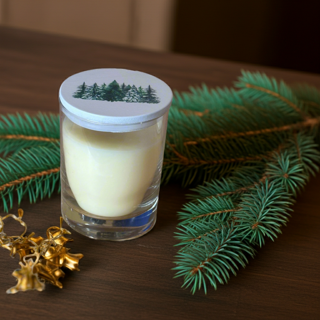 Candle in a glass jar with a forest-themed lid on a wooden surface with greenery and gold decorations.